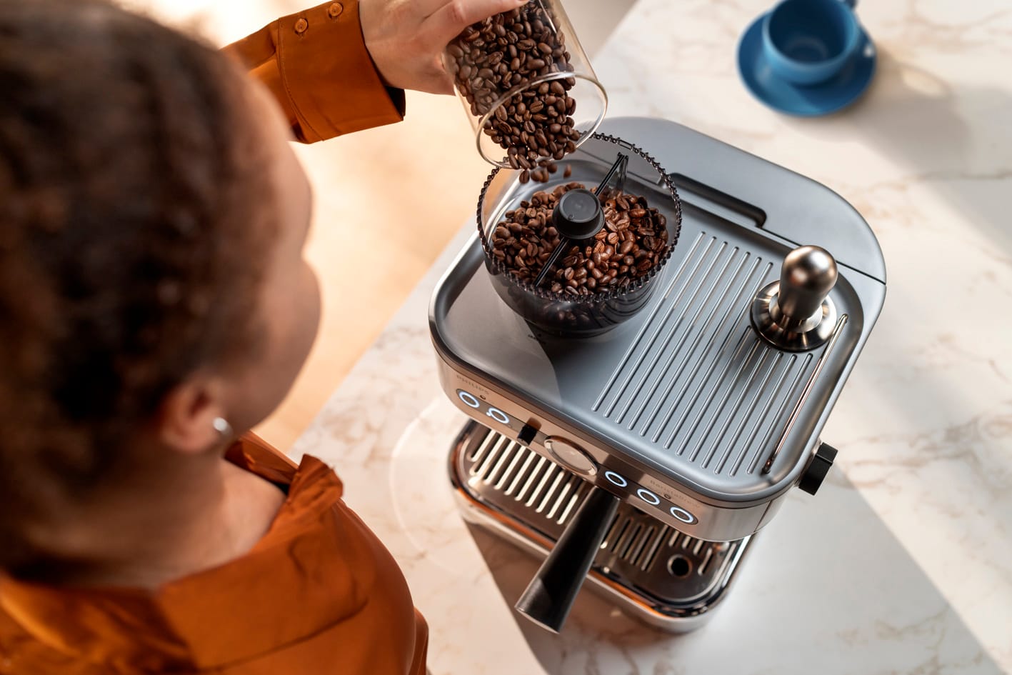 Mlad&aacute; žena sype zrnkovou k&aacute;vu do n&aacute;doby na k&aacute;vov&aacute; zrna k&aacute;vovaru Philips Barista Brew